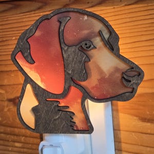 May include: A wooden night light featuring a dog's head silhouette. The dog's profile is filled with a sunset scene of orange, red, and yellow hues. The night light is set in a white plug-in base.