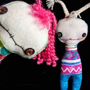 K&ouml;nnte beinhalten: Zwei handgefertigte Stoffpuppen mit gestickten Gesichtsz&uuml;gen und Knopfaugen. Eine Puppe hat pinkfarbenes Garnhaar und ein buntes Patchworkkleid. Die andere Puppe hat wei&szlig;es Garnhaar und ein lila, pink und blaues gestreiftes Kleid.