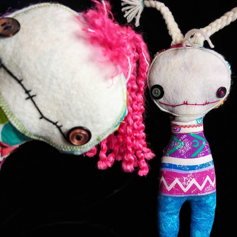 K&ouml;nnte beinhalten: Zwei handgefertigte Stoffpuppen mit gestickten Gesichtsz&uuml;gen und Knopfaugen. Eine Puppe hat pinkfarbenes Garnhaar und ein buntes Patchworkkleid. Die andere Puppe hat wei&szlig;es Garnhaar und ein lila, pink und blaues gestreiftes Kleid.