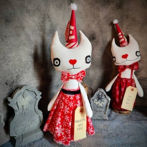 May include: Two white fabric cat dolls wearing red and white Christmas outfits. The dolls are standing on a shelf with a gray background. One doll has a tag that says "Naughty and Nice". The other doll has a tag that says "Stay Scary".