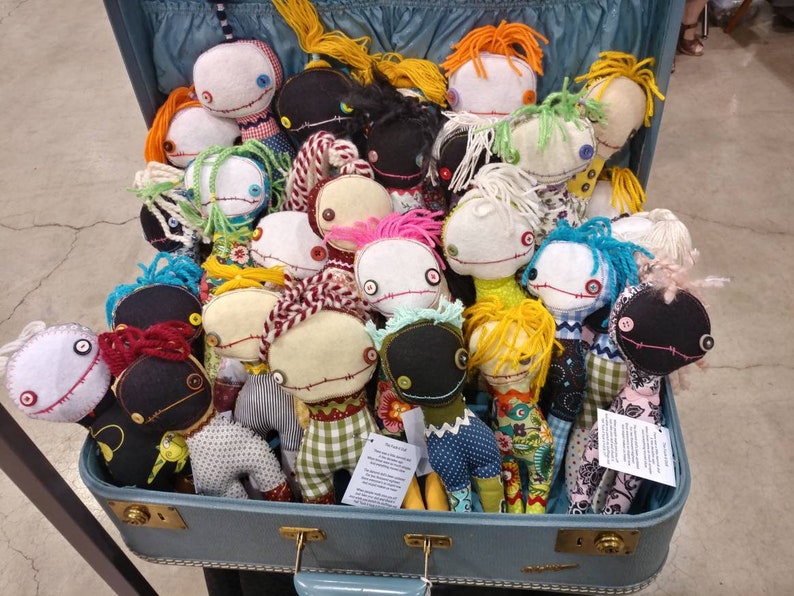 K&ouml;nnte beinhalten: Ein Vintage-Koffer gef&uuml;llt mit handgefertigten Stoffpuppen. Die Puppen sind aus verschiedenen Stoffen gefertigt und haben unterschiedlich farbiges Garnhaar. Einige Puppen haben Kn&ouml;pfe als Augen, andere haben gestickte Augen. Die Puppen sind alle einzigartig und haben einen skurrilen Charme.