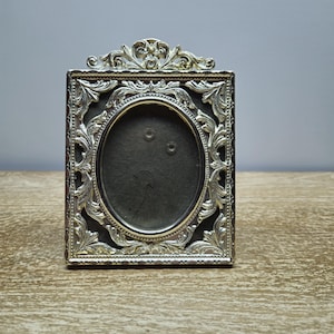 Può includere: Cornice per foto ornata argentata con un'apertura ovale. La cornice presenta un design decorativo con dettagli intricati.