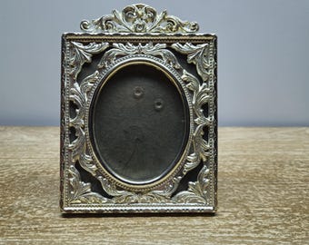 May include: Ornate silver-toned picture frame with an oval opening. The frame features a decorative design with intricate details.