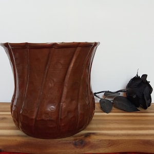 Pode incluir: Um vaso de cobre marrom com acabamento martelado e um design ondulado. O vaso está sobre uma superfície de madeira.