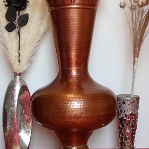 Peut inclure: Un grand vase en cuivre martelé avec une large ouverture évasée et une base ronde. Le vase est de couleur brun rouge foncé.