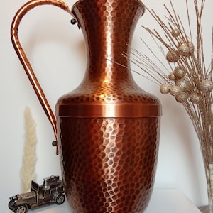 May include: A hammered copper vase with a wide mouth and a handle. The vase has a textured surface and a warm, reddish-brown colour.