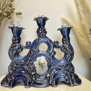 Può includere: Un candelabro in ceramica blu e bianco con tre portacandele, dettagli a volute ornamentali e un medaglione ovale centrale decorato con un piccolo bouquet floreale. L'oggetto ha una finitura lucida con accenti in argento metallizzato.