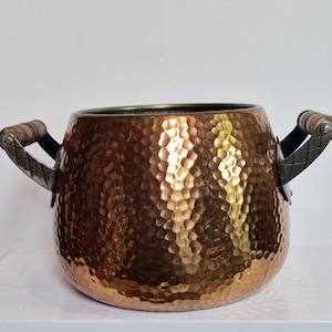 Op de afbeelding: Een koperkleurige gehamerde metalen kom met twee handvatten. De handvatten zijn gemaakt van donker metaal met een houten handvat. De kom heeft een getextureerd oppervlak en een ronde vorm. De kom wordt waarschijnlijk gebruikt voor decoratieve doeleinden.