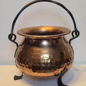 Könnte beinhalten: Ein Kupferkessel mit gehammerter Textur, mit einem schwarzen Metallgriff und drei kleinen Füßen. Der Kessel hat einen abgerundeten Körper und einen breiten Rand. Der Griff ist gebogen und die Füße sind stilisiert.