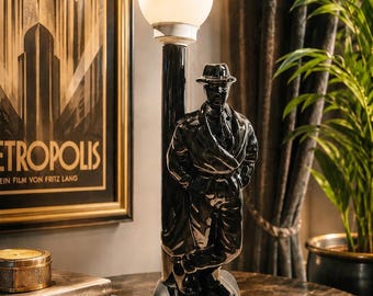 Op de afbeelding: Een zwarte Art Deco lamp met een figuur van een man met hoed en jas, leunend tegen een zwarte paal met een wit gloeilamp. De lamp staat op een tafel met marmeren blad, met een ingelijste "Metropolis" filmposter op de achtergrond.