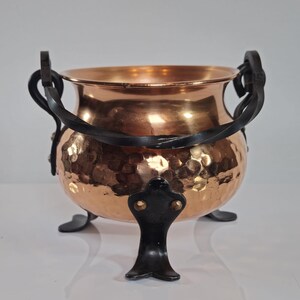 Könnte beinhalten: Ein Kupferkessel mit gehämmertem Muster, getragen von drei schwarzen Füßen. Der Kessel hat zwei schwarze Griffe und eine dekorative schwarze Halterung. Der Kupfertopf hat eine warme, metallische Farbe.