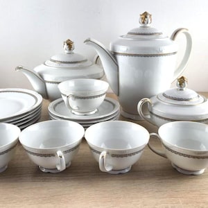 Puede incluir: Un juego de té de porcelana blanca que incluye una tetera alta, un azucarero con tapa, una tetera con tapa, varios platillos y varias tazas de té. Cada pieza está decorada con una banda fina de patrón dorado y negro y remates dorados.
