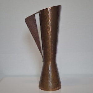 Op de afbeelding: Een hoge, gehamerde koperen vaas met een houten handvat. De vaas heeft een uitlopende bovenkant en een smalle basis. Het houten handvat is aan de zijkant van de vaas bevestigd. De vaas heeft een warme, metallic bruine kleur.