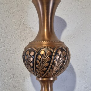 Könnte beinhalten: Eine bronzefarbene dekorative Vase mit einem breiten, bauchigen Körper und einem schlanken Hals. Die Vase weist aufwendige Blatt- und seilartige Muster um den Körper auf. Der Sockel ist als Sockel gestaltet, und der Gesamtstil ist verziert und elegant.
