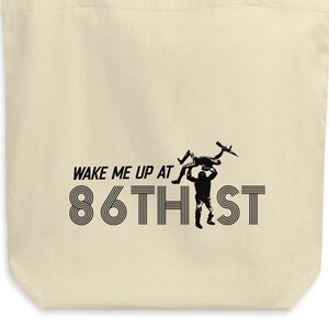 May include: A beige tote bag with black text that reads "Wake me up at 86th St." and a graphic of a person carrying a drone.