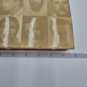 May include: A vintage book with a red spine and a beige cover featuring a grid of nine black and white portraits. The book is measured with a ruler, showing a length of approximately 6 inches.