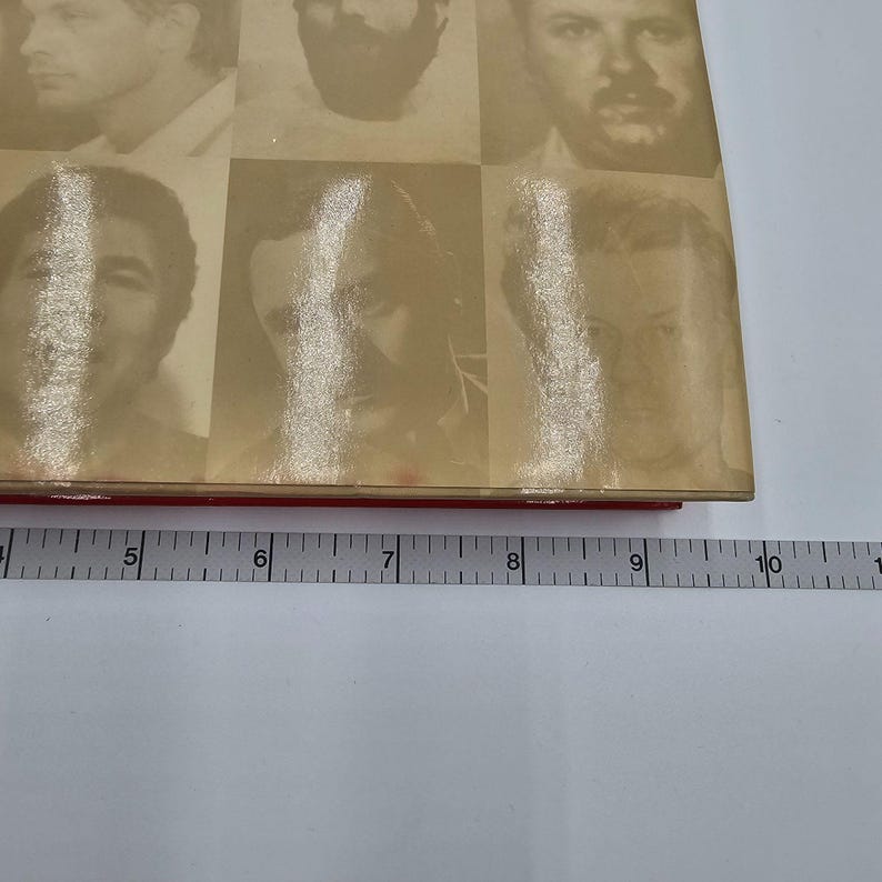 May include: A vintage book with a red spine and a beige cover featuring a grid of nine black and white portraits. The book is measured with a ruler, showing a length of approximately 6 inches.