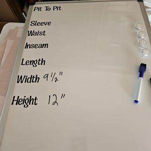 May include: Whiteboard with handwritten measurements in black marker. The measurements include: Width 9 1/2", Height 12". Other labels include: Pit To Pit, Sleeve, Waist, Inseam, and Length. A blue and white dry-erase marker is visible.
