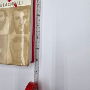May include: A book titled "BLACKHALL" with a sepia-toned cover featuring four men's faces. A red measuring tape is positioned next to the book, showing measurements in inches.