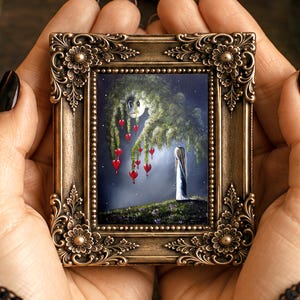 Puede incluir: Un peque&ntilde;o marco rectangular con un borde ornamentado de color bronce que presenta tallas florales. El marco contiene una pintura de una figura de pie sobre una colina cubierta de hierba bajo un &aacute;rbol llor&oacute;n con adornos rojos en forma de coraz&oacute;n.