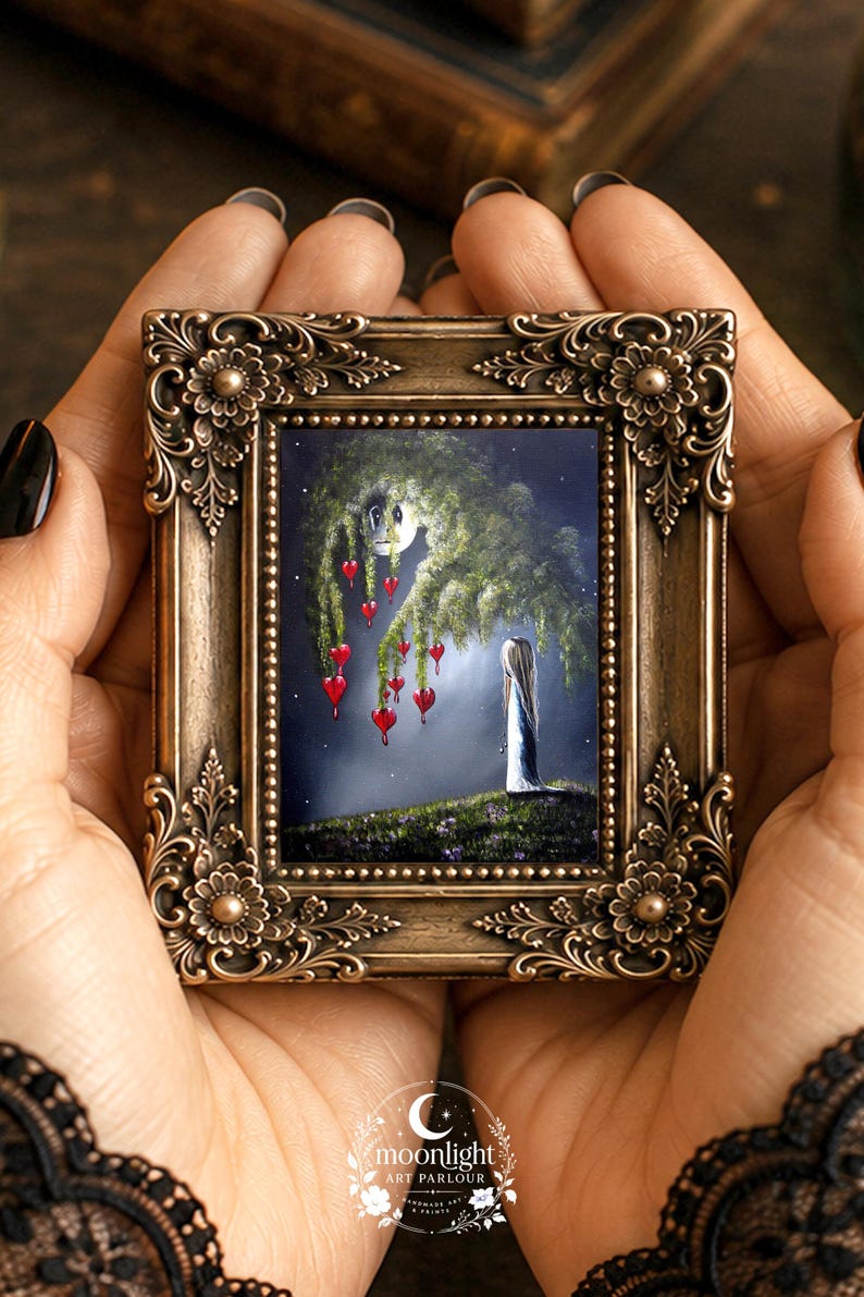 Puede incluir: Un peque&ntilde;o marco rectangular con un borde ornamentado de color bronce que presenta tallas florales. El marco contiene una pintura de una figura de pie sobre una colina cubierta de hierba bajo un &aacute;rbol llor&oacute;n con adornos rojos en forma de coraz&oacute;n.