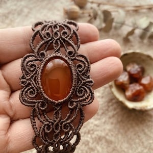 May include: A brown macrame bracelet featuring a central oval-shaped smooth orange carnelian gemstone. The bracelet is crafted from dark brown cord tied in intricate, symmetrical knotted patterns that surround the stone and form the band.