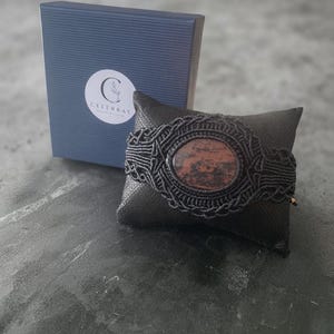 May include: A black macrame bracelet featuring a central oval-shaped reddish-brown stone with dark inclusions. The bracelet is displayed on a small black pillow next to a blue gift box with a white circular label that reads CELEBRAY.
