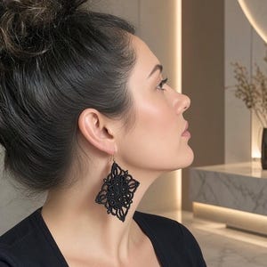 Op de afbeelding: Een zwarte macramé oorbel met een ingewikkeld, ruitvormig ontwerp van geknoopte koorden. De oorbel hangt aan een eenvoudige goudkleurige metalen haak en toont een gedetailleerd, symmetrisch patroon van lussen en geweven texturen in een donkere tint.
