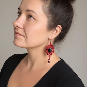 May include: A long macrame earring featuring a central oval black stone surrounded by intricate red cord knotting. The design includes a smaller black bead dangling at the bottom, suspended from a dark metal hook, creating a detailed drop earring.
