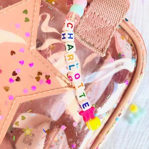 May include: A pink, clear, star-shaped bag with a beaded charm that spells out the name "CHARLOTTE". The charm features colorful beads and star-shaped accents. The bag is decorated with confetti and has a pink strap.
