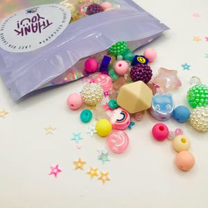 May include: A clear plastic bag filled with colourful beads, including stars, smiley faces, and rainbow shapes. The bag is labelled "THANK YOU!" and has a German phrase on it.