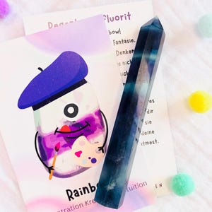 Op de afbeelding: Een blauw en wit fluoriet kristalpunt staat naast een kaart met een cartoon illustratie van een verfpot met een baret. De kaart heeft het woord "Rainbow" en andere tekst. Kleurrijke pompons zijn verspreid.