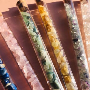 Könnte beinhalten: Sechs Reagenzgläser aus Glas, gefüllt mit verschiedenen farbigen Kristallen. Die Kristalle sind klein und scheinen geschliffene Steine zu sein. Die Reagenzgläser sind aus klarem Glas mit Korkstopfen.
