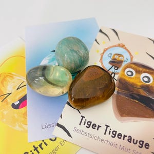 Könnte beinhalten: Drei polierte Trommelsteine in Braun, durchscheinendem Gelb und Hellgrün auf drei illustrierten Karten. Die Karten zeigen Cartoon-Grafiken und deutschen Text, darunter Tiger Tigerauge und Citrin, sowie Muster mit Tigerstreifen.