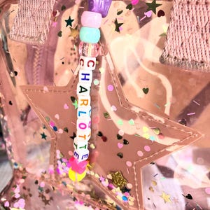 May include: A clear pink backpack with a star-shaped pocket and a beaded charm. The charm spells out the name "CHARLOTTE" with colourful beads. The backpack is decorated with star and heart-shaped confetti.