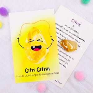 Könnte beinhalten: Eine gelbe Karte mit einer Cartoon-Zitrin-Illustration und dem Text "Citri Citrin". Ein kleiner Citrin-Stein und eine Karte mit deutschem Text sind ebenfalls vorhanden. Rosa und lila Pompons sind verstreut.