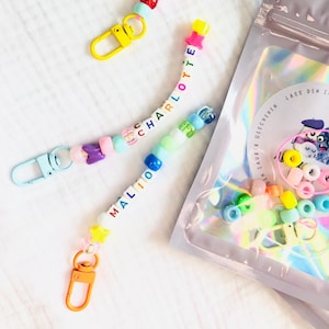 May include: Handmade beaded keychains spelling out "CHARLOTTE" and "MALIO" in colourful letter beads. Each keychain has a different coloured clasp. A bag of beads and accessories is visible in the background.
