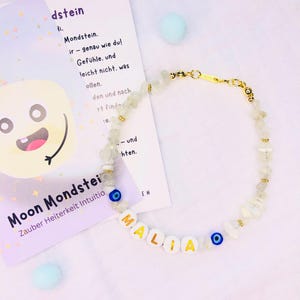 Könnte beinhalten: Ein handgefertigtes Armband mit dem Namen "MALIA" in goldenen Buchstaben auf weißen Perlen. Das Armband hat klare, unregelmäßig geformte Steine und goldfarbene Akzente. Zwei blaue Evil-Eye-Perlen sind in das Design integriert. Im Hintergrund liegt eine Karte mit deutschem Text.