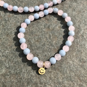 Può includere: Un set di bracciale e collana di perline con perline alternate di pietre preziose azzurre e rosa. Il bracciale ha un ciondolo color oro con una faccina sorridente. I gioielli sono esposti su una superficie grigia strutturata.