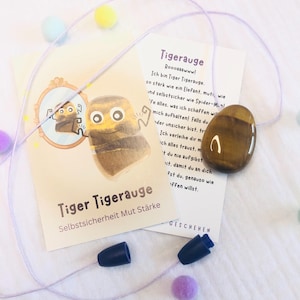 Könnte beinhalten: Ein Tigeraugen-Edelsteinanhänger an einer lila Kordel, mit einer Karte mit einer Cartoon-Tiger-Illustration und deutschem Text. Die Karte trägt auch die Worte "Tiger Tigerauge" und "Selbstsicherheit Mut Stärke". Bunte Pompons sind verstreut.