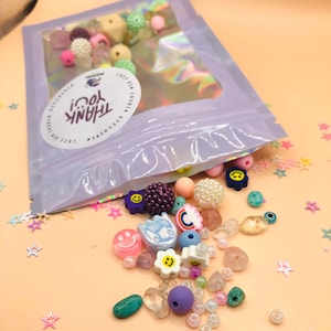 Puede incluir: Una bolsa morada con una ventana transparente llena de cuentas de colores, dijes y otros suministros para hacer joyas. La bolsa está abierta y el contenido se derrama sobre una superficie naranja claro. La bolsa tiene el texto "THANK YOU!" impreso en ella.