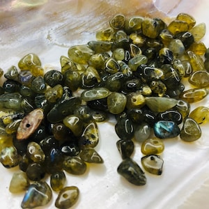 Pode incluir: Uma coleção de pequenas pedras de labradorite polidas em tons de verde, cinza e dourado. As pedras estão espalhadas sobre uma superfície branca e iridescente, criando uma estética natural. Algumas pedras exibem flashes azuis e iridescentes.