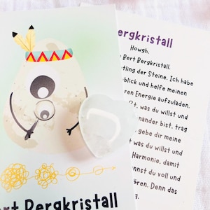 Op de afbeelding: Een helder kwartskristal in de vorm van een hart ligt op een wit oppervlak naast een kaart met Duitse tekst en een cartoonillustratie. De kaart toont het woord "ergkristall".