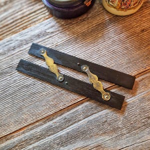 May include: Two rectangular dark wood strips featuring decorative brass hardware. Each strip has a scalloped brass plate secured with small rivets. The wood has a visible grain texture and the brass components show a metallic finish with subtle patina.