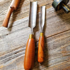 May include: Two wood carving chisels with metal blades and wooden handles, along with a wooden marking gauge and a wooden handle, are arranged on a weathered wooden surface. The chisels have brass ferrules.