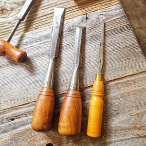 May include: Three woodworking chisels with polished metal blades and smooth, rounded wooden handles. Two of the chisels feature a letter D stamped on the metal ferrule, while the third, thinner tool has a lighter wood handle and a slender blade.