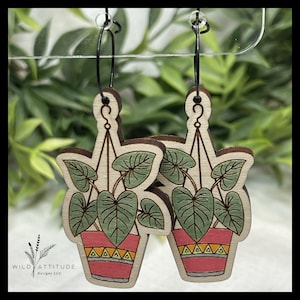 May include: Wooden earrings featuring a plant in a hanging pot design. The earrings have a light wood base with a detailed illustration of a plant with green leaves and a pink pot with yellow and orange accents. Black metal hoops.