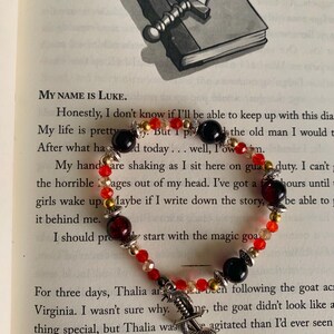 May include: A beaded bracelet with red, black, and gold beads, and a silver dagger charm. The bracelet is on a book page with text. A silver dagger rests on a closed book.