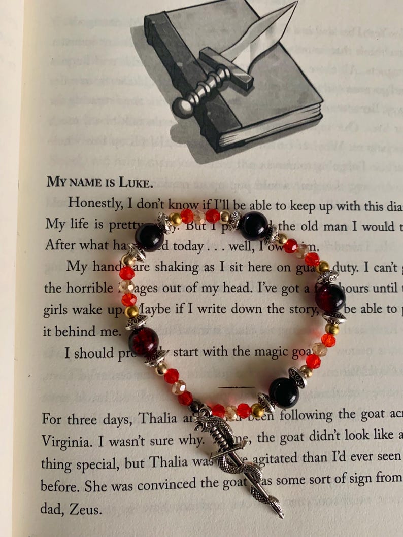 May include: A beaded bracelet with red, black, and gold beads, and a silver dagger charm. The bracelet is on a book page with text. A silver dagger rests on a closed book.