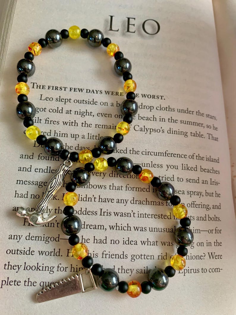 May include: Two beaded bracelets with black, yellow, and orange beads. One bracelet has a silver charm of a hammer, and the other has a saw charm. The word "LEO" is visible in the background.